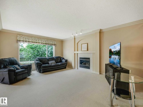 Living room featuring a gas fireplace with a white mantel and arched alcove, track lighting, crown molding, and a large window overlooking mature greenery - 1116 Lincoln Crescent, Edmonton, AB - Indoor Photo Showing Living Room With Fireplace