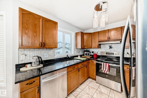 U-shaped kitchen featuring wood-finish cabinetry, dark countertops, a subway tile backsplash, stainless steel appliances, and a window above the sink - 8 Campbell Court, Leduc, AB 