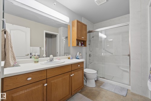 Bathroom featuring a wood-finish vanity with an integrated sink, a large wall-mounted mirror, and a full shower enclosure with a clear glass door - 413 8912 156 Street Nw, Edmonton, AB 