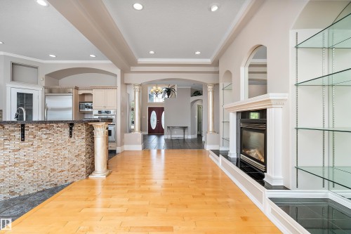 Open-concept living area featuring light wood-finish flooring, a built-in fireplace with a black surround, integrated glass shelving, and decorative architectural columns - 16103 89 Street, Edmonton, AB - Indoor With Fireplace