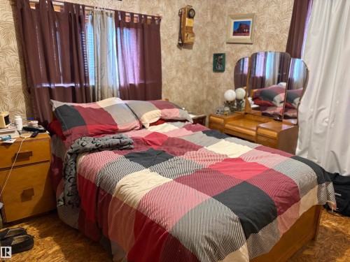 Bedroom featuring patterned wallpaper, a window with sheer and opaque curtains, a wooden nightstand, and a wooden dressing table with an attached tri-fold mirror. - 125 56524 Rng Rd 52A, Lindbergh, AB 