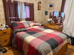 Bedroom featuring patterned wallpaper, a window with sheer and opaque curtains, a wooden nightstand, and a wooden dressing table with an attached tri-fold mirror. - 