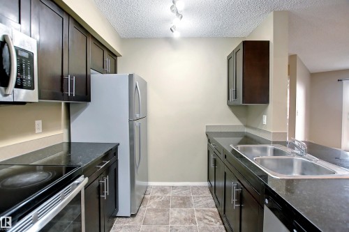Galley kitchen featuring dark wood cabinetry, stainless steel appliances including a microwave and double-door refrigerator, dark stone countertops, a dual-basin stainless steel sink, and tile flooring - 215 1510 Watt Drive, Edmonton, AB 