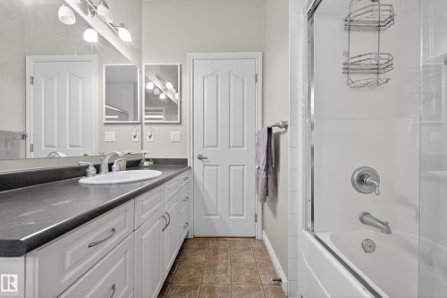 Spacious bathroom featuring a single vanity with an integrated sink and dark countertop, white cabinetry with brushed nickel hardware, a full wall mirror, and a shower-over-tub combination with a glass enclosure - 4113 59 Street, Beaumont, AB 