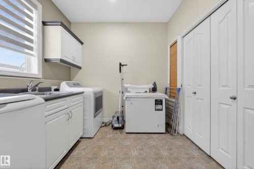 Dedicated laundry room featuring a utility sink, dark countertop, white cabinetry with silver hardware, and tiled flooring - 4113 59 Street, Beaumont, AB 