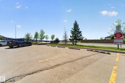 Paved parking lot featuring designated parking spaces with wheel stops, surrounded by mature trees and residential structures in the background - 225 1510 Watt Drive, Edmonton, AB - Outdoor