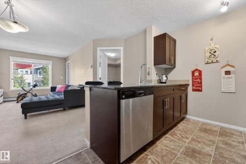 Contemporary kitchen area featuring wood-finish cabinetry, stainless steel dishwasher, and a dark stone countertop - 225 1510 Watt Drive, Edmonton, AB - Indoor