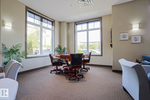 Spacious room featuring broad windows with roller shades, neutral tone walls, patterned carpet flooring, and integrated wall sconces - 117 160 Magrath Road, Edmonton, AB - Indoor Photo Showing Office
