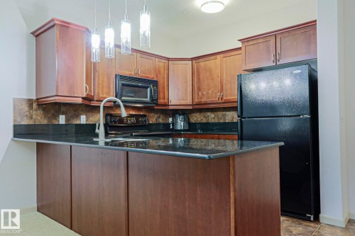Kitchen with wood-finish cabinetry, black appliances, dark countertops, and a tiled backsplash - 117 160 Magrath Road, Edmonton, AB - Indoor Photo Showing Kitchen