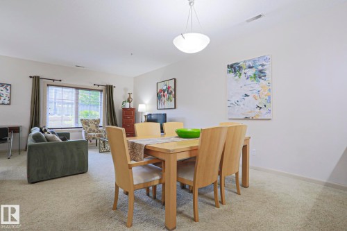 Carpeted dining area with a contemporary chandelier, featuring an adjacent living space with a large window and blinds - 117 160 Magrath Road, Edmonton, AB - Indoor Photo Showing Other Room