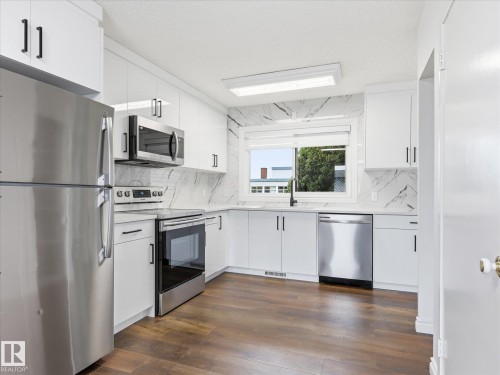 Modern kitchen featuring white cabinetry, stainless steel appliances, marble-look backsplash, wood-finish flooring, and a window above the sink - 13316 136 Avenue, Edmonton, AB 