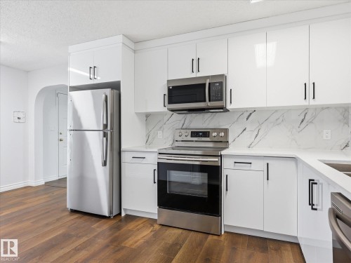 Kitchen featuring white cabinetry with black hardware, stainless steel appliances, a marble-look backsplash, and wood-finish flooring - 13316 136 Avenue, Edmonton, AB 