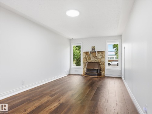 Spacious room featuring wood-finish flooring, a stone-clad fireplace with metal insert, and two large windows - 13316 136 Avenue, Edmonton, AB 