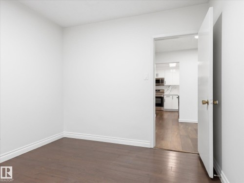 Bright room featuring dark wood-finish flooring, white baseboards, and a doorway opening to a kitchen with white cabinetry, stainless steel appliances, and a marble-patterned backsplash - 13316 136 Avenue, Edmonton, AB 