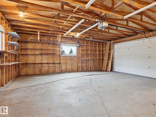 Spacious garage featuring exposed wood framing, concrete flooring, built-in shelving, two windows, and an overhead garage door with an automatic opener - 13316 136 Avenue, Edmonton, AB 