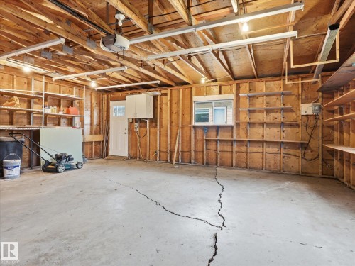 Spacious garage featuring exposed wood framing, concrete flooring, built-in shelving, an interior access door, and multiple overhead lights - 13316 136 Avenue, Edmonton, AB 