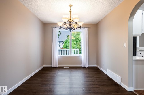 Spacious room featuring dark wood-finish flooring, a contemporary ceiling chandelier, a large window with white trim, and an arched entry - 16811 91 Street, Edmonton, AB 