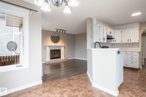 Open-concept kitchen and living area featuring a fireplace with a white mantel, wood-finish flooring, white cabinetry with dark hardware, and a tile backsplash - 16811 91 Street, Edmonton, AB 