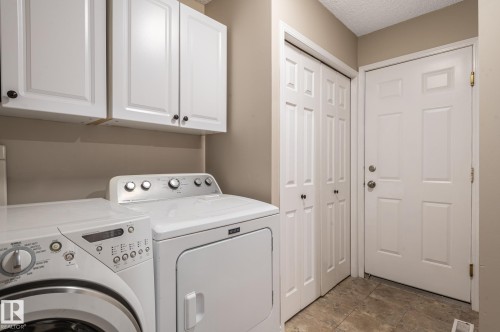 Laundry area featuring overhead white cabinetry with dark hardware, bi-fold closet doors, a paneled door, and neutral-toned floor tiling - 16811 91 Street, Edmonton, AB 
