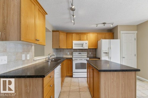 Kitchen featuring wood-finish cabinetry, dark countertops, a tiled backsplash, a central island, and white appliances - 12215 18 Avenue Sw, Edmonton, AB 