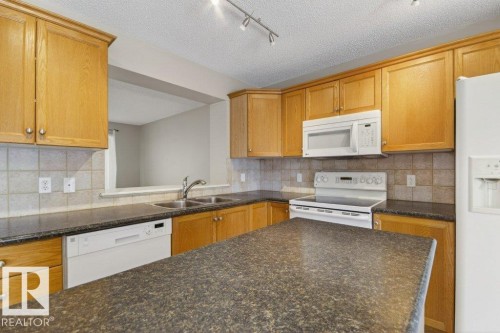 Kitchen featuring ample wood cabinetry, speckled countertops, a tile backsplash, and white appliances including a built-in microwave and dishwasher - 12215 18 Avenue Sw, Edmonton, AB 