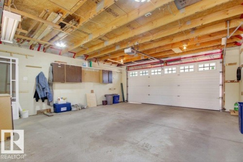 Spacious two-car garage featuring a sectional overhead door with window inserts, exposed wood ceiling joists, concrete flooring, and built-in wall cabinets - 12215 18 Avenue Sw, Edmonton, AB 