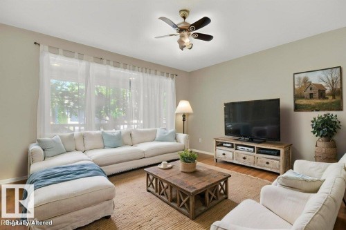 Spacious living area featuring large windows with sheer curtain treatments, wood-finish flooring, a ceiling fan with integrated lighting, and neutral wall tones. Virtually staged - 12215 18 Avenue Sw, Edmonton, AB 