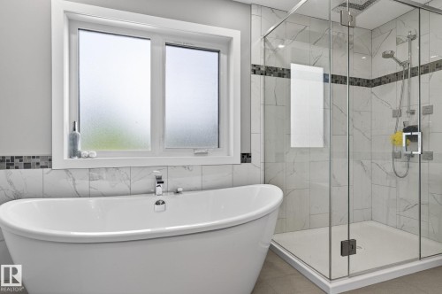 Spacious bathroom featuring a freestanding soaking tub with deck-mounted chrome fixtures, a glass-enclosed shower with multiple showerheads, white-veined tile walls, and a decorative mosaic accent band - 8809 148 Street, Edmonton, AB - Indoor Photo Showing Bathroom