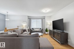 Bright living area featuring wood-finish flooring, light grey painted walls, a window with curtains, a modern ceiling light fixture, and a wooden staircase railing - 