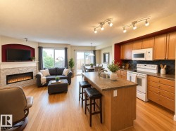 Open-concept living space featuring wood-finish flooring, a kitchen island with seating, a stacked stone fireplace, a sliding glass door to the exterior, and track lighting - 