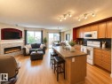 Open-concept living space featuring wood-finish flooring, a kitchen island with seating, a stacked stone fireplace, a sliding glass door to the exterior, and track lighting - 102 Hartwick Loop, Spruce Grove, AB 