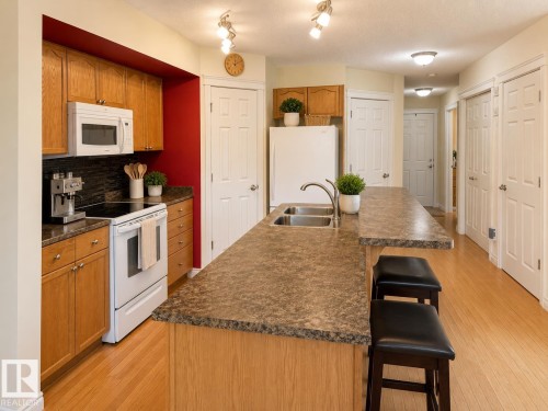 Functional kitchen featuring an eat-at island, dual-basin stainless steel sink, wood-finish cabinetry, built-in microwave, and wood-finish flooring - 102 Hartwick Loop, Spruce Grove, AB 