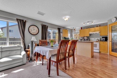 Open-concept living area featuring a dining space with wood-finish flooring, a kitchen with light wood cabinetry and white appliances, and a carpeted seating area with large windows - 14031 136 Street, Edmonton, AB 