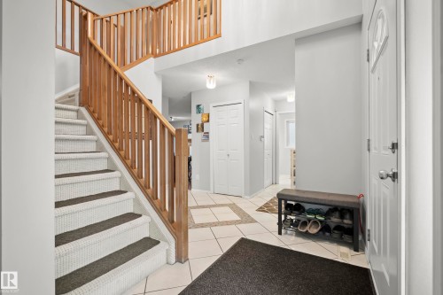 Foyer featuring a two-story entryway, wood balusters, carpeted staircase with dark treads, light tile flooring with a contrasting tile inlay, and a white front entry door with an arched window insert - 15503 62 Street, Edmonton, AB 