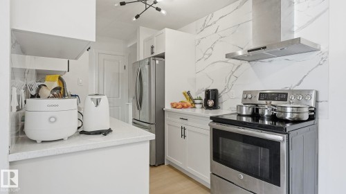 Kitchen featuring a full-height marble-patterned backsplash, stainless steel range with overhead hood, integrated stainless steel refrigerator, white cabinetry with dark hardware, and light-toned countertops - 20C Callingwood Court, Edmonton, AB - Indoor Photo Showing Kitchen