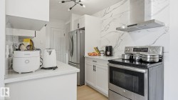 Kitchen featuring a full-height marble-patterned backsplash, stainless steel range with overhead hood, integrated stainless steel refrigerator, white cabinetry with dark hardware, and light-toned countertops - 