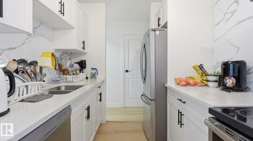 Galley kitchen featuring white cabinetry with black hardware, light-toned countertops, and a white marble-look backsplash - 20C Callingwood Court, Edmonton, AB - Indoor Photo Showing Kitchen With Double Sink
