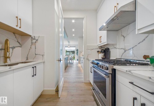Galley kitchen featuring white cabinetry with gold and black hardware, stone countertops with integrated sink, stainless steel range with hood, and wood-finish flooring - 16607 8 Street, Edmonton, AB 