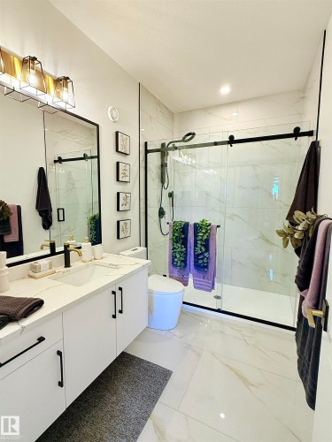 Modern bathroom featuring a vanity with a white countertop and rectangular sink, black-framed mirror, contemporary lighting fixture, glass-enclosed shower with black trim, and large format tiled flooring and shower surround - 16607 8 Street, Edmonton, AB 