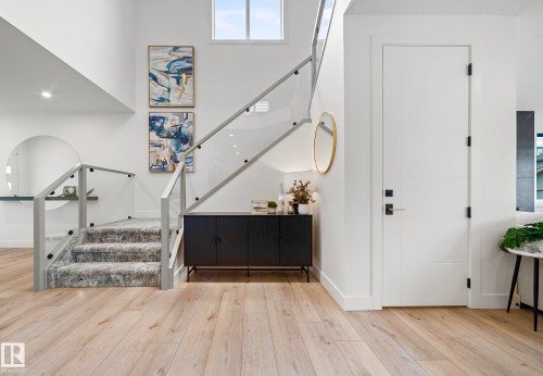 Bright entryway featuring light wood-finish flooring, a modern glass-paneled staircase with carpeted treads, a white paneled door with black hardware, and high ceilings with upper windows - 16607 8 Street, Edmonton, AB 