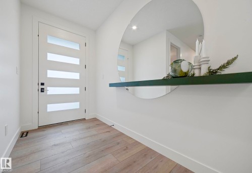 Entryway featuring a contemporary white door with frosted glass panels, wood-finish flooring, a wall-mounted circular mirror, a green floating shelf, and recessed lighting - 16607 8 Street, Edmonton, AB 