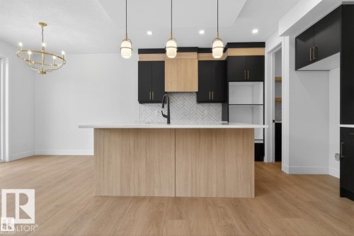 Open-concept kitchen featuring a light wood-finish island with a white countertop, contrasting black and natural wood cabinetry, herringbone tile backsplash, and three pendant lights - 19 Rosa Crescent, St. Albert, AB 