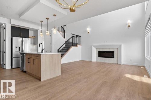 Spacious open-concept interior featuring light wood-finish flooring, a contemporary kitchen island with a white countertop and dark cabinetry, and a black metal staircase with wood treads - 19 Rosa Crescent, St. Albert, AB 