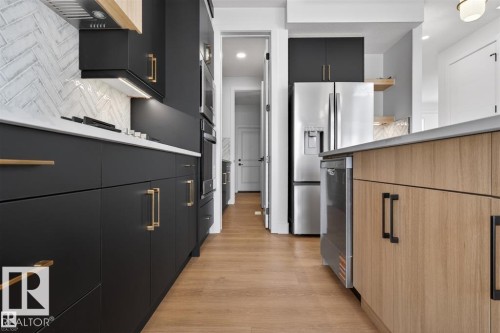 Modern kitchen featuring matte black cabinetry with gold-tone hardware, wood-finish island cabinetry, stainless steel appliances, a herringbone tile backsplash, and wood-finish flooring - 19 Rosa Crescent, St. Albert, AB 