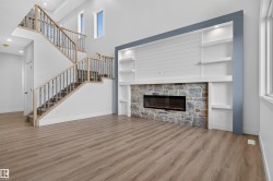 Open-concept living area featuring wood-finish flooring, a contemporary fireplace with a stone surround, built-in shelving, a modern staircase with wrought iron balusters, and abundant natural light from multiple windows - 
