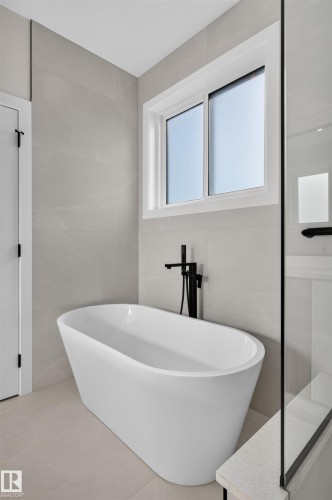 Freestanding soaking tub with a matte black floor-mounted faucet, complemented by light-toned wall and floor tiling - 84 Rosa Crescent, St. Albert, AB - Indoor Photo Showing Bathroom