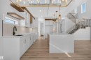 Open-concept living space with light wood-finish flooring, a two-story staircase with dark metal balusters, a large kitchen island with a light-toned countertop, white cabinetry with gold-tone hardware, and multiple modern pendant light fixtures - 84 Rosa Crescent, St. Albert, AB  - Indoor 