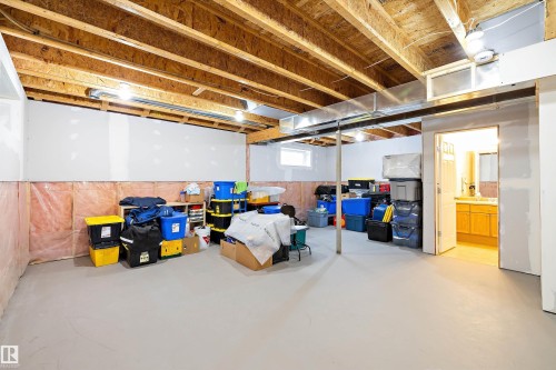 Spacious unfinished basement with exposed wood joists and concrete flooring, featuring a partially finished wall, a small window, and an open doorway revealing a vanity with wood cabinetry - 3344 24 Avenue, Edmonton, AB 