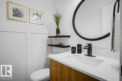 Bathroom featuring a wood-finish vanity with a white countertop and integrated sink, matte black faucet, and a large round mirror with a textured frame - 