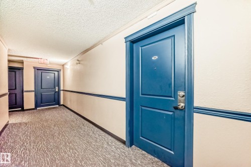 Corridor featuring textured carpeting, neutral wall finishes, and contrasting blue doors with trim - 108 10535 122 Street, Edmonton, AB 